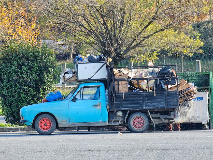 A small blue pickup truck parked on the side of a street, loaded with an overflow of various types of rubbish and waste materials. The truck's bed and rear section are filled with flattened cardboard 