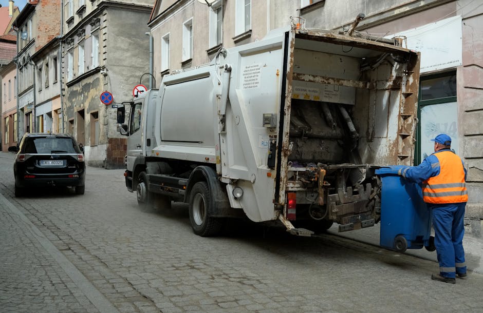 Two large black trash bags filled with waste materials are resting against a black metal fence on the side of a street pavement. The bags appear tightly packed and are made of glossy plastic with refl