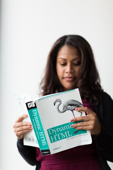 A woman with medium-length dark hair, dressed in a black jacket and maroon top, is reading a book titled 'Dynamic HTML' with a cover featuring an illustration of a heron. She is standing indoors again